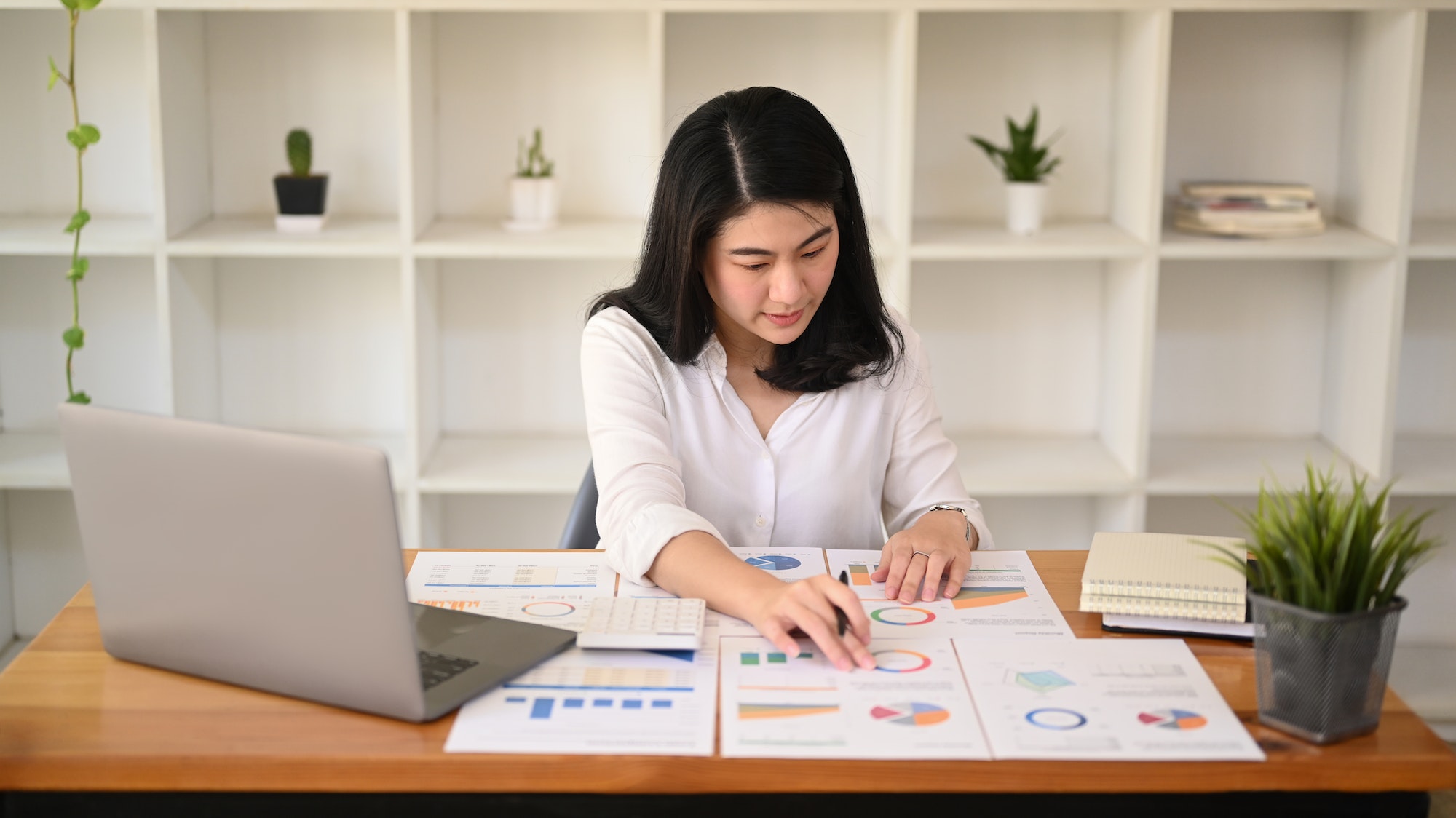 Concentrated asian woman accountant using laptop and analyzing financial graph on wooden table.